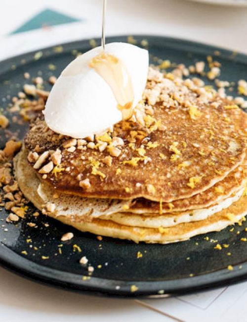 A stack of pancakes garnished with nuts and a dollop of cream on a black plate, with syrup being poured on top.
