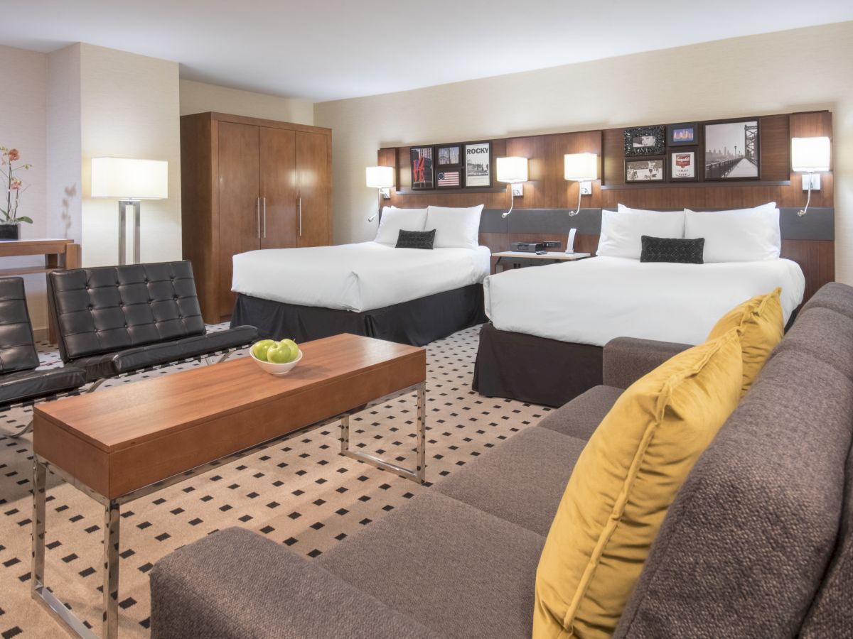 A modern hotel room with two double beds, a sofa, a long wooden coffee table, two lounge chairs, and a wooden wardrobe in the background.