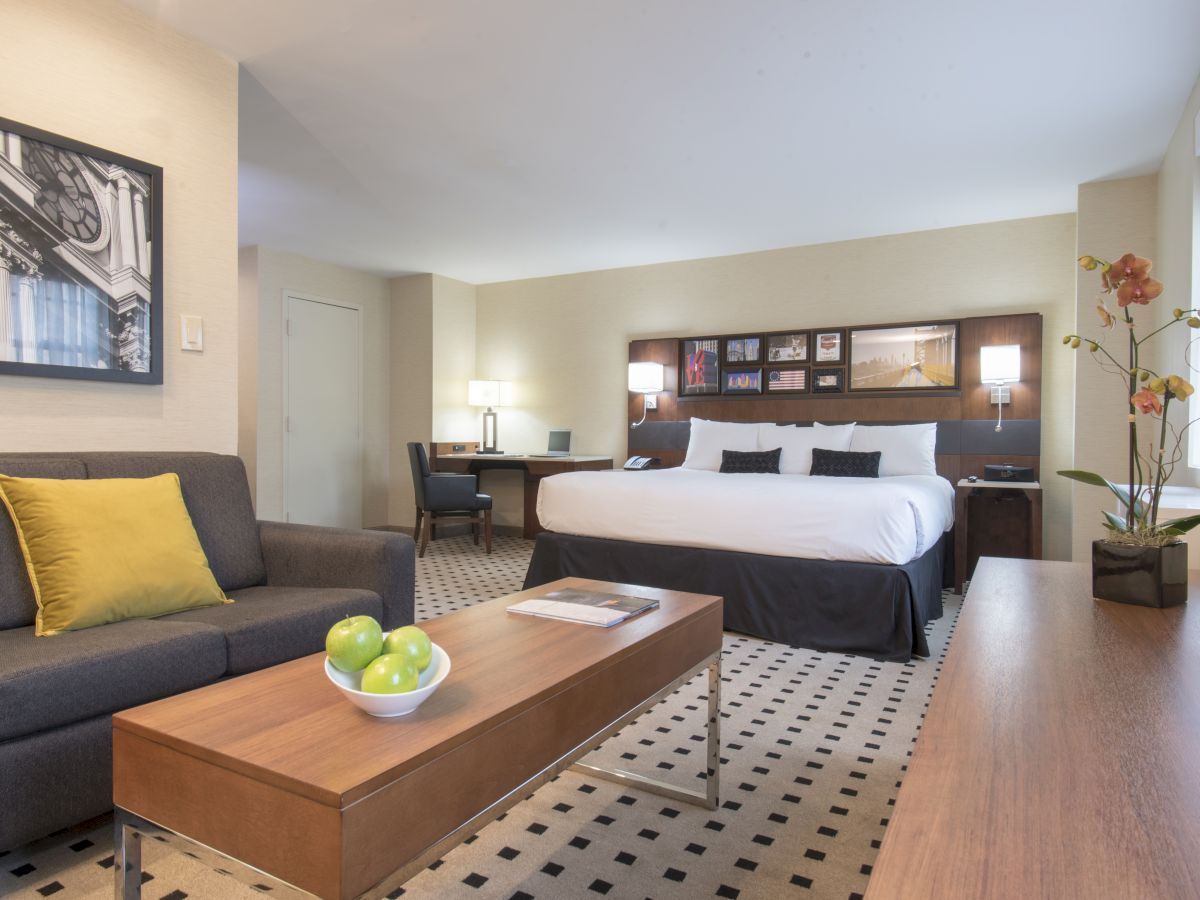 A modern hotel room with a king-sized bed, a gray sofa, a wooden coffee table with green apples, a desk, and contemporary wall art.