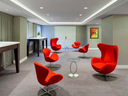 A modern room with six red chairs, small round tables, and white walls with artwork, featuring a minimalist design and a gray carpet.