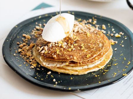 A stack of pancakes topped with whipped cream and syrup on a black plate. Crushed nuts are sprinkled around the pancakes.