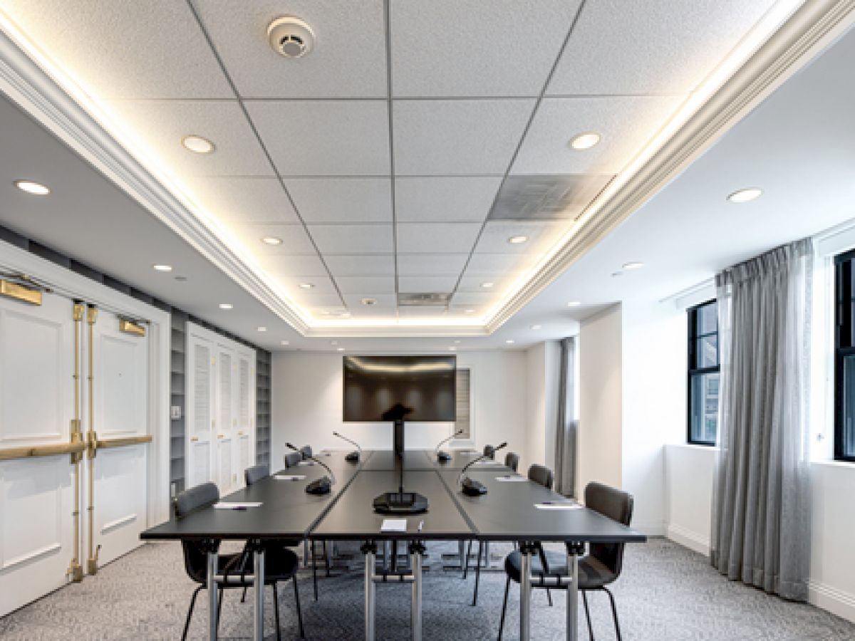 A modern conference room with a long table, chairs, microphones, a large screen, and windows with curtains. The ceiling has recessed lighting.