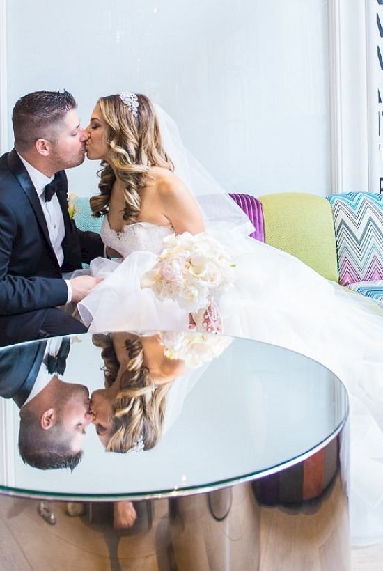 A couple in wedding attire shares a kiss in a modern room with colorful seating and mirrored table, reflecting them. Text on the wall is partially visible.