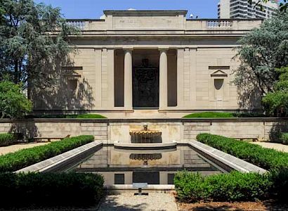 A classical building fronted by a symmetrical garden with a rectangular reflective pool, trimmed hedges, and a decorative central fountain.