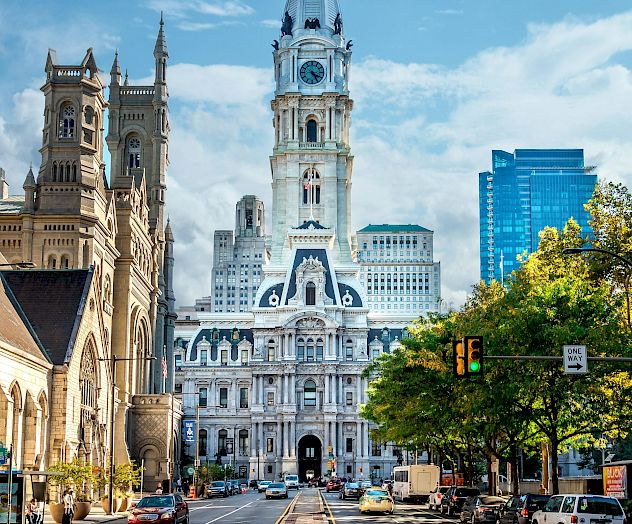 A view of a city street leading to a grand historic building with a tall clock tower, surrounded by other buildings and a blue sky with clouds.